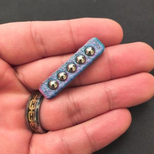 Puede incluir: Un objeto pequeño, rectangular, azul y rosa con cinco rodamientos de bolas de metal plateado. El objeto se sostiene en la mano de una persona, con un anillo plateado y dorado en el dedo.