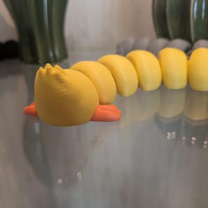 May include: A yellow articulated caterpillar toy with a rounded head, small orange feet, and a segmented body. The toy features a smooth, matte finish and is constructed from multiple connected pieces that allow for flexible movement and posing.