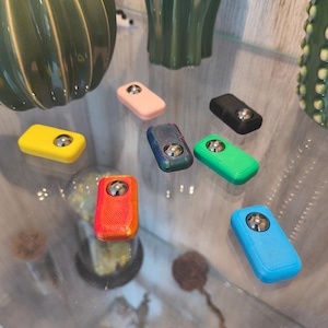 May include: A collection of colorful fidget toys in various shades, including yellow, pink, blue, green, and red. Each rectangular toy features a silver ball bearing in the center. The toys are displayed on a glass surface.