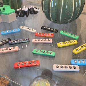 Puede incluir: Una colección de juguetes magnéticos rectangulares y coloridos. Cada juguete tiene un color diferente, incluyendo azul, rosa, verde, amarillo y rojo. Cada rectángulo tiene seis rodamientos de bolas de metal plateado. Los juguetes están dispuestos sobre una superficie de vidrio.