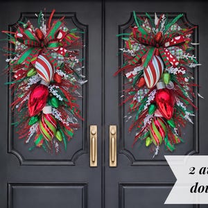 May include: Two teardrop-shaped door swags featuring red, green, and white striped ornaments, large red bulb-shaped decorations, plaid ribbons, and white frosted greenery. The text 2 available for double doors is printed on a white banner.