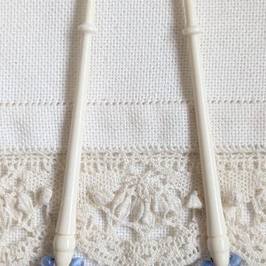 May include: Two ivory-coloured bobbin lace tools with blue and white bead accents. The tools are set against a white fabric background with a decorative lace trim. These tools are used for bobbin lace making.