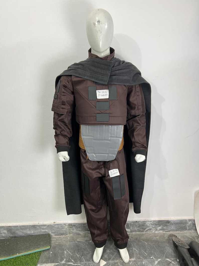 May include: A brown costume with a gray cape and accents. The costume includes a jacket, pants, and a chest piece. The text "The Deal Digger" is visible on the costume. The mannequin is wearing white shoes.