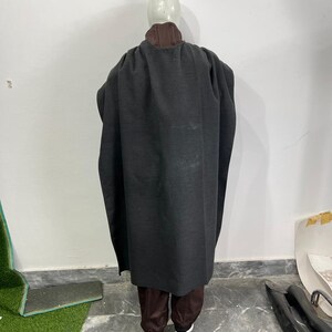 May include: A dark gray cloak with a brown collar and matching brown pants. The cloak is long and flowing, covering the mannequin's body. The pants are a darker shade of brown. The mannequin is wearing white shoes.