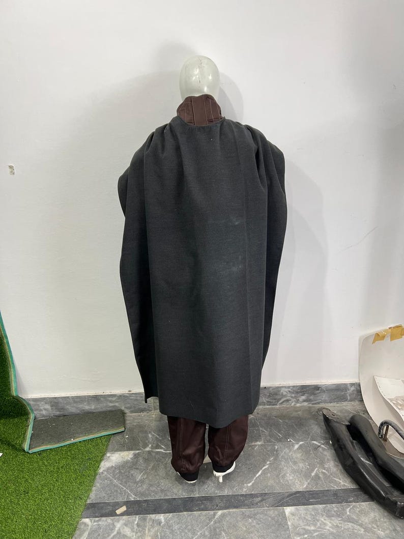 May include: A dark gray cloak with a brown collar and matching brown pants. The cloak is long and flowing, covering the mannequin's body. The pants are a darker shade of brown. The mannequin is wearing white shoes.