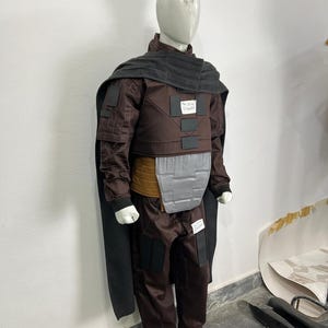 May include: A brown costume with a black cape and gray accents is displayed on a mannequin. The costume includes a jacket, pants, and a chest plate. The mannequin is wearing white shoes.