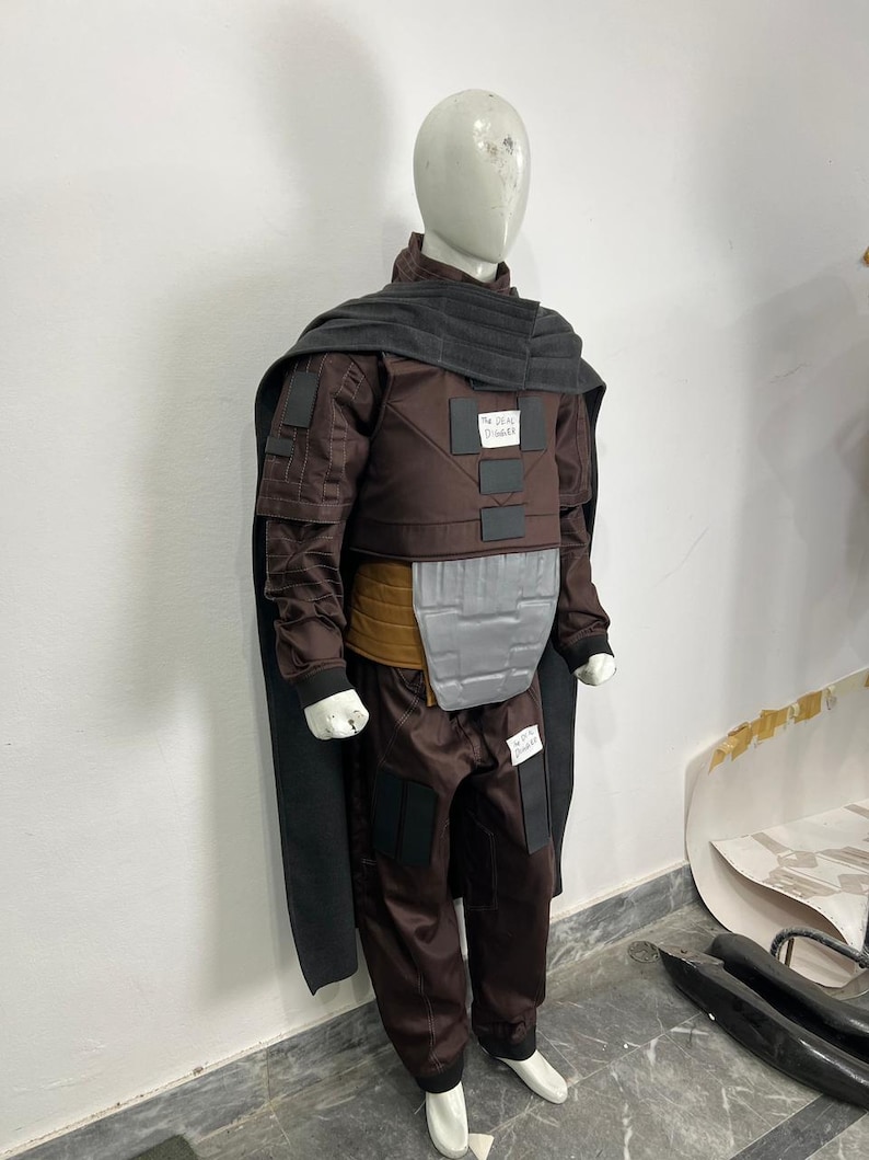 May include: A brown costume with a black cape and gray accents is displayed on a mannequin. The costume includes a jacket, pants, and a chest plate. The mannequin is wearing white shoes.