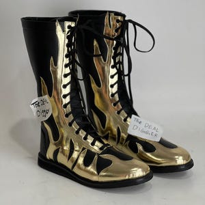 Pode incluir: Um par de botas de luta livre pretas e douradas com desenhos de chamas. As botas com atacadores são feitas de materiais brilhantes. A etiqueta "The Deal Digger" está presa a cada bota. Estas botas são projetadas para luta livre profissional.