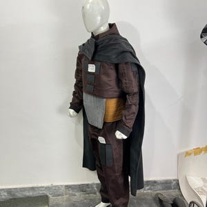 May include: A brown and black costume displayed on a mannequin. The costume includes a jacket, pants, and a cape. The jacket has a gray chest piece and a tan belt. The pants have black pockets and white shoes.