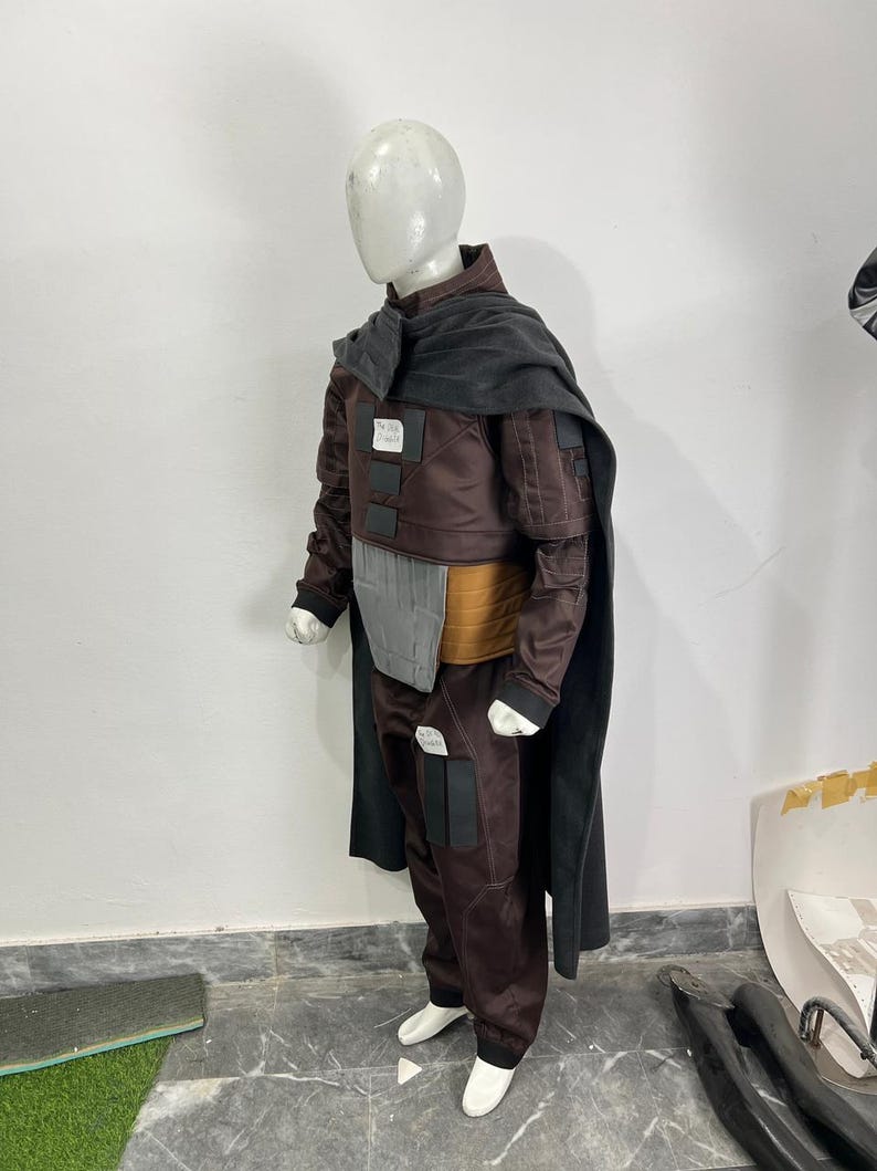 May include: A brown and black costume displayed on a mannequin. The costume includes a jacket, pants, and a cape. The jacket has a gray chest piece and a tan belt. The pants have black pockets and white shoes.
