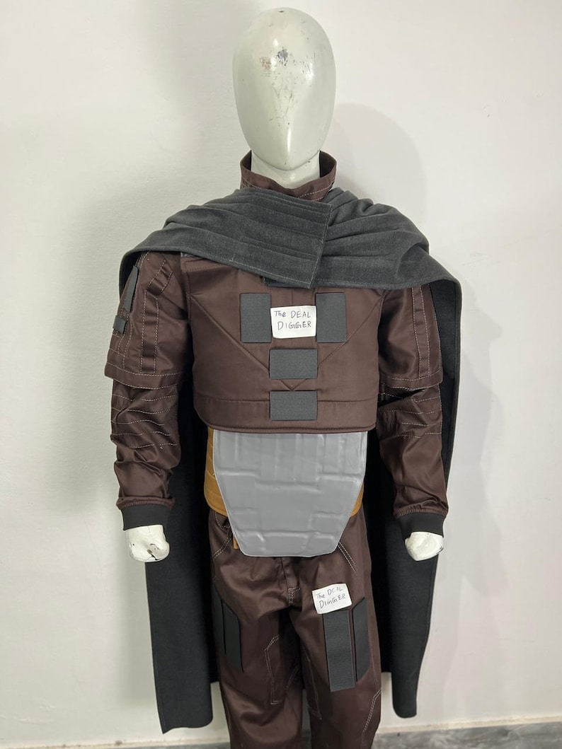 May include: A brown costume with a gray cape and accents, displayed on a mannequin. The costume includes a jacket, pants, and a chest plate. The text "THE DEAL DIGGER" is visible on the costume.