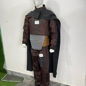May include: A brown costume with a gray chest plate, a dark gray cape, and matching pants. The costume includes a brown jacket with the text "The Real Disaster" on a patch. The mannequin is wearing white shoes.