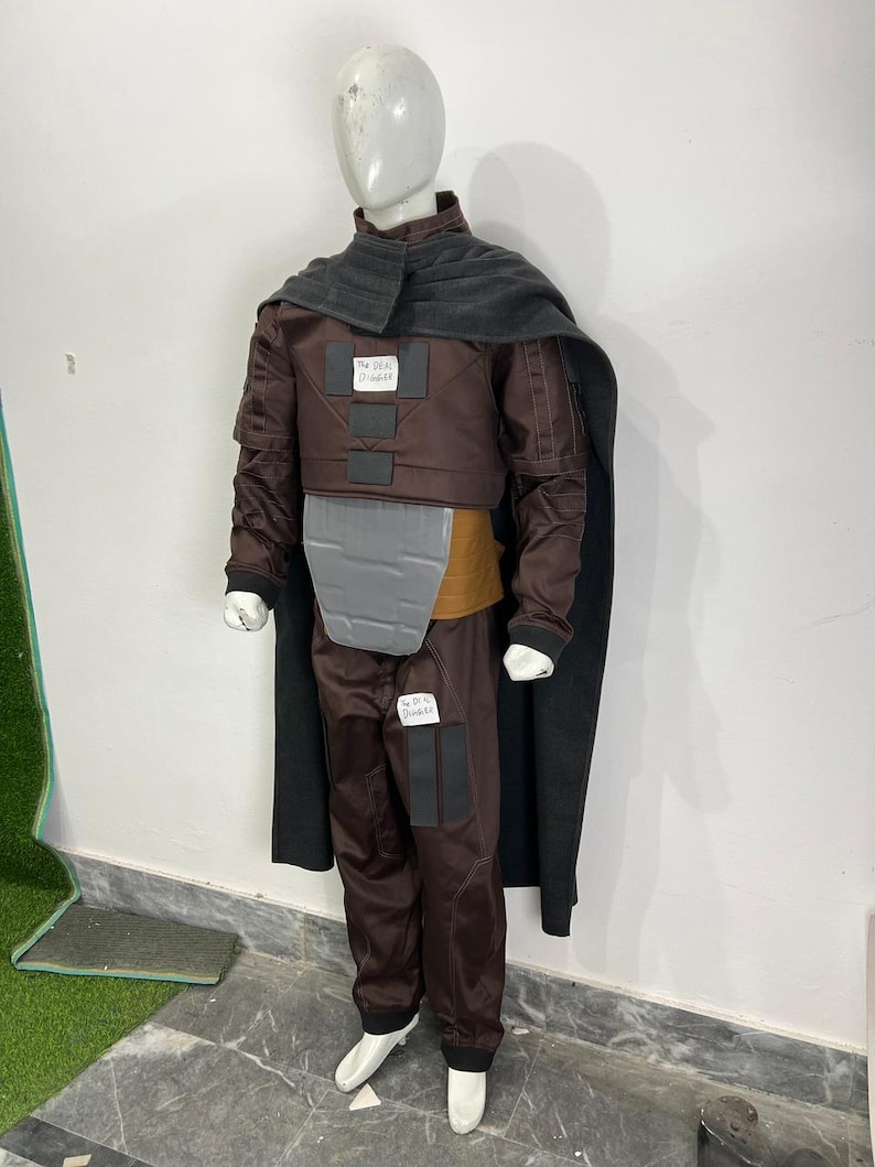 May include: A brown costume with a gray chest plate, a dark gray cape, and matching pants. The costume includes a brown jacket with the text "The Real Disaster" on a patch. The mannequin is wearing white shoes.