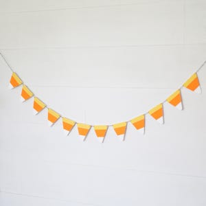 May include: A festive garland featuring candy corn-shaped pennants. Each pennant has a white, yellow, and orange color scheme, strung together on a silver chain. The garland is displayed against a white background, perfect for Halloween or fall decor.
