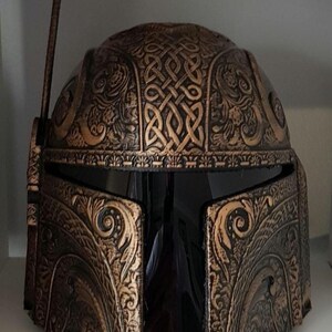 Op de afbeelding: Een zwarte en gouden helm met ingewikkelde decoratieve details en een kleine antenne bovenop. De helm heeft een donker vizier en is gemaakt van een getextureerd materiaal.