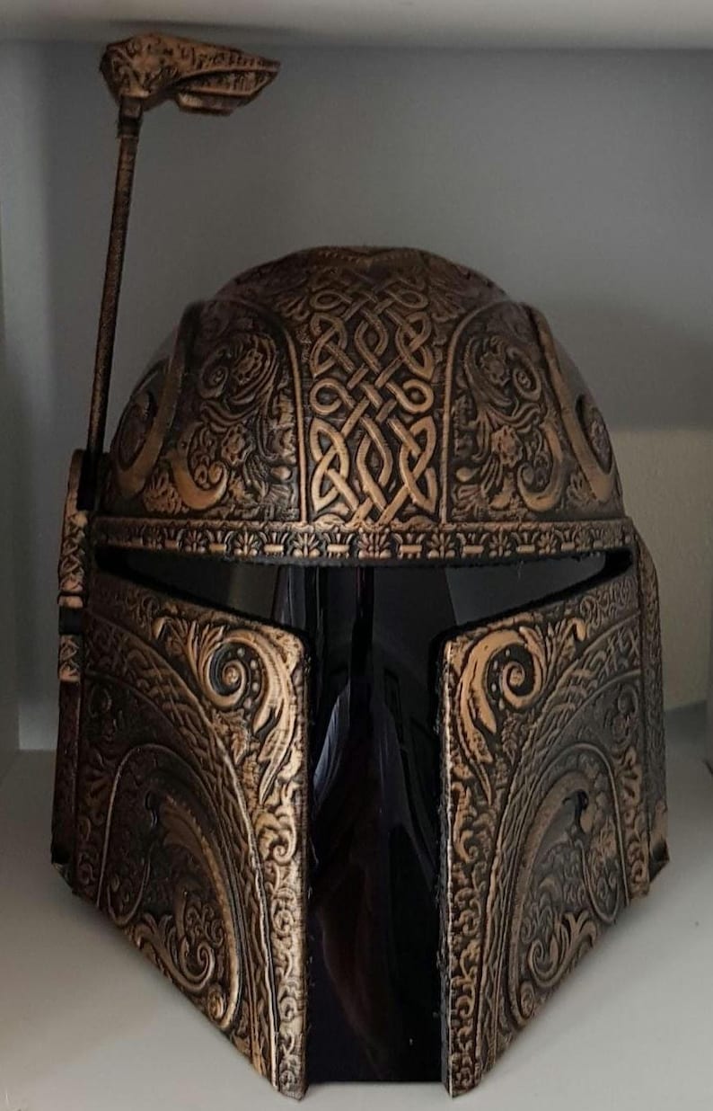 Op de afbeelding: Een zwarte en gouden helm met ingewikkelde decoratieve details en een kleine antenne bovenop. De helm heeft een donker vizier en is gemaakt van een getextureerd materiaal.