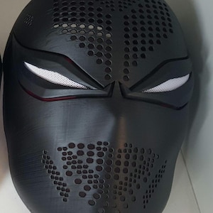 May include: A matte black full-face mask featuring a textured surface with numerous small circular perforations across the forehead and mouth areas. The mask includes two narrow, angular eye openings covered with a fine white mesh material.