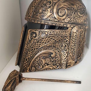 Op de afbeelding: Een gouden en zwarte helm met ingewikkelde bloem- en Keltische knoopontwerpen. De helm is gemaakt van een hard materiaal en heeft een vizier. De helm is een rekwisiet voor cosplay of kostuum.