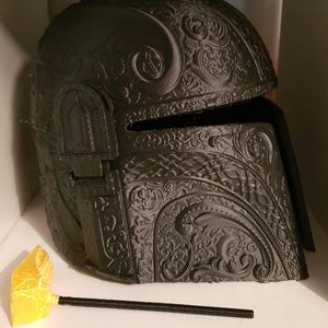 Op de afbeelding: Een zwarte helm met ingewikkelde, sierlijke ontwerpen. De helm is gemaakt van een 3D-geprint materiaal en heeft een vizier.