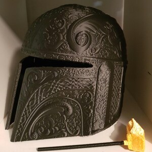 Op de afbeelding: Een zwarte 3D-geprinte helm met ingewikkelde bloem- en geometrische ontwerpen. De helm heeft een vizier en een puntige bovenkant.