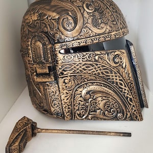 Op de afbeelding: Een gouden en zwarte helm met ingewikkelde bloem- en Keltische knoopontwerpen. De helm heeft een vizier en een afneembare antenne.