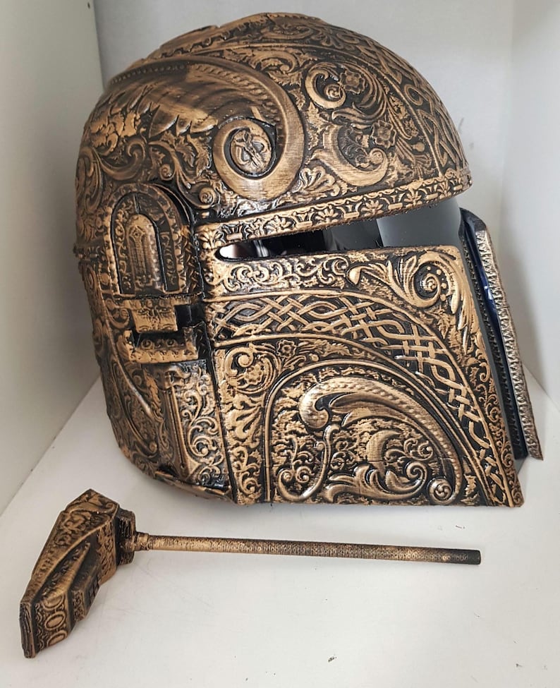 Op de afbeelding: Een gouden en zwarte helm met ingewikkelde bloem- en Keltische knoopontwerpen. De helm heeft een vizier en een afneembare antenne.