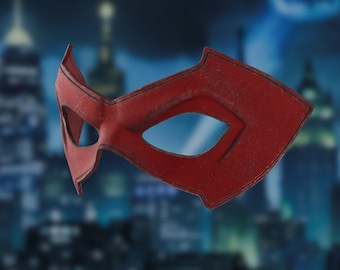 May include: A red domino mask featuring a textured surface and black stitched detailing along the edges. The mask has two large, angular eye openings and a pointed design that extends outward at the temples, creating a sharp, defined silhouette.