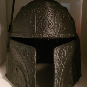 Op de afbeelding: Een zwarte plastic helm met ingewikkelde, gedetailleerde gravures. De helm heeft een verhoogde, gele, plastic antenne bovenop.