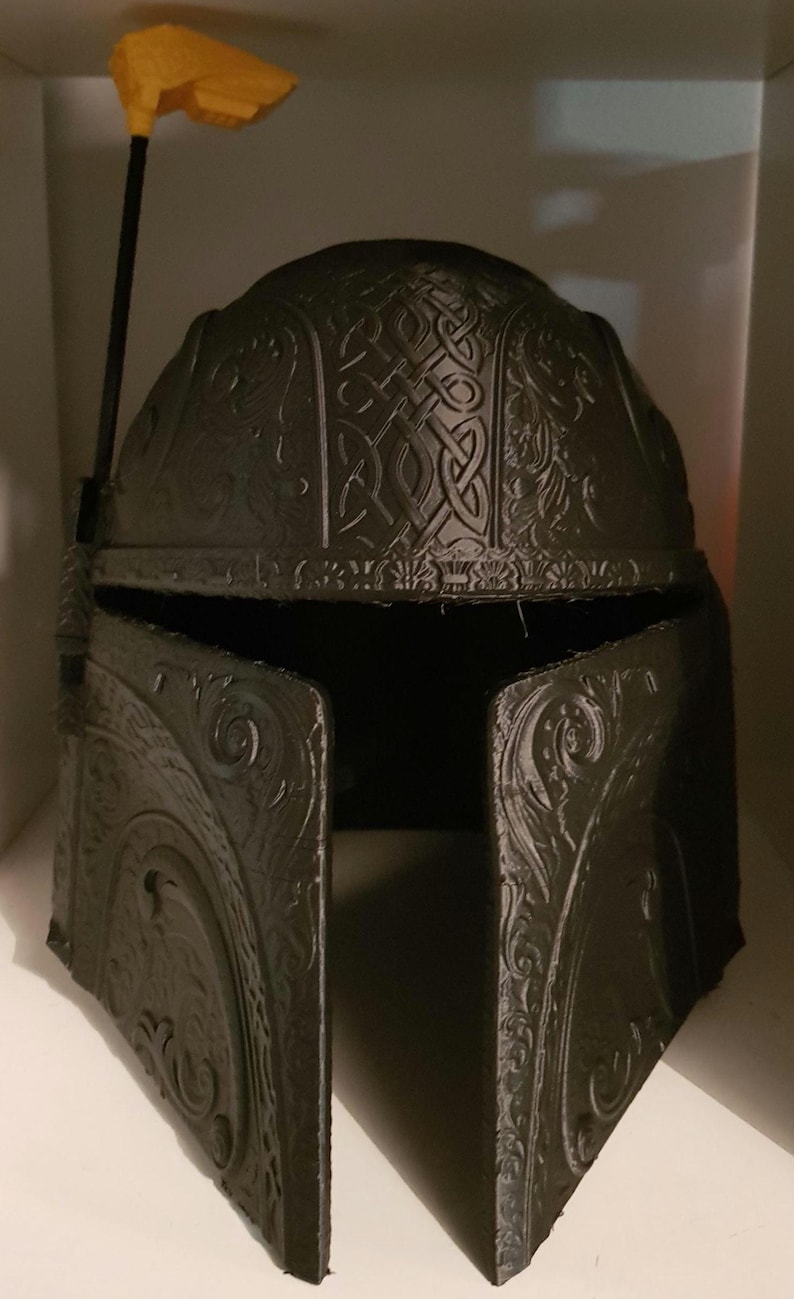 Op de afbeelding: Een zwarte plastic helm met ingewikkelde, gedetailleerde gravures. De helm heeft een verhoogde, gele, plastic antenne bovenop.
