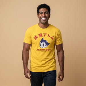 May include: A mustard yellow short-sleeve t-shirt featuring a graphic of the Astro Boy character in blue and white. Above the character is the red Japanese text 鉄腕アトム, and below is the red text ASTRO BOY and the smaller text SINCE 1952.