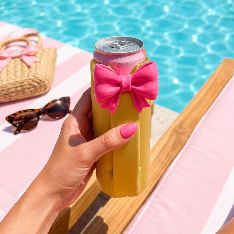 May include: A yellow can cooler with a bright pink bow is held in front of a pink can. The background includes a pool, sunglasses, and a straw bag with a pink bow. The scene is set on a pink and white striped surface.