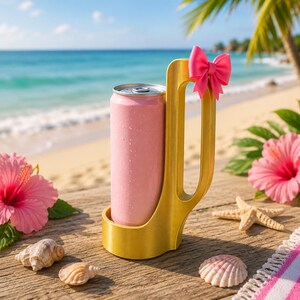 May include: A pink beverage can in a gold-colored holder with a pink bow, set on a wooden surface with seashells and hibiscus flowers. The background features a beach and ocean scene.