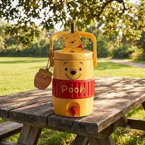 May include: A yellow beverage cooler featuring a Winnie the Pooh face, a red band with the text Pooh, a brown straw, a handle, and a hanging charm shaped like a honey pot with the word HUNNY printed on it, including a red spigot at the base.