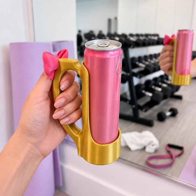 May include: A pink beverage can held in a gold-colored handle with a pink bow. The handle is designed to hold the can securely. The background includes gym equipment, suggesting a fitness or active lifestyle theme.