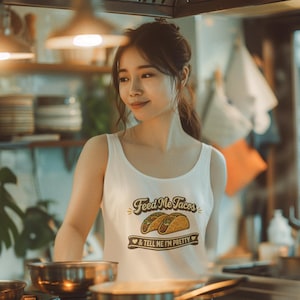 以下が含まれることがあります： 「Feed Me Tacos & Tell Me I'm Pretty」の文字がプリントされた白いタンクトップ。キッチンで調理器具や鍋を背景に、人が写っています。温かい照明です。