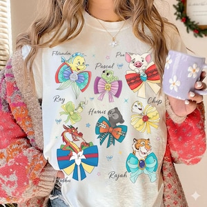 Op de afbeelding: Een wit t-shirt met kleurrijke cartoonkarakterillustraties, elk met een strik en een naam zoals Flounder, Pascal en Pua. Het shirt heeft een ronde hals en korte mouwen. De persoon houdt een paarse mok met witte bloemmotieven vast.