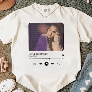May include: White t-shirt with a graphic of a music player interface displaying a photo of a woman. The text on the graphic reads "Silent Treatment" by Freya Skye. The shirt has a silver chain around the neckline.