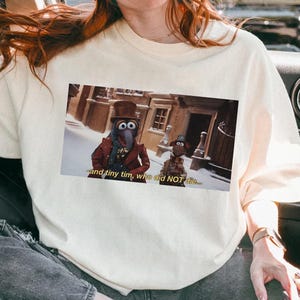 Puede incluir: Camiseta color crema con una imagen rectangular de dos personajes de los Muppets de una escena de una película. La imagen incluye el texto "and tiny tim, who did NOT die...". La camiseta es una prenda informal para el día a día.