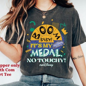 May include: Dark gray t-shirt with a colorful graphic design. The design includes the text "OO BABY! IT'S MY MEDAL. NO TOUCHY!" and "KUZCOTOPIA VIRTUAL 5K" with a "runDisney" logo. The shirt is worn by a person.
