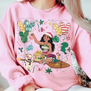 K&ouml;nnte beinhalten: Rosafarbenes Sweatshirt mit einer Cartoon-Illustration einer Frau mit Weihnachtsm&uuml;tze, umgeben von festlichen Elementen. Mickey Mouse-f&ouml;rmige Luftballons, ein Schwein und ein Hahn sind ebenfalls abgebildet. Das Design hat ein Feiertags-Thema.