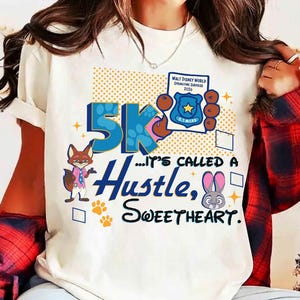 Könnte beinhalten: Cremefarbenes T-Shirt mit einem Grafikdesign mit dem Text "5K...IT'S CALLED A Hustle, SWEETHEART." und Zeichentrickfiguren. Das Design enthält ein Abzeichen mit der Aufschrift "Walt Disney World Springtime Surprise 2026".