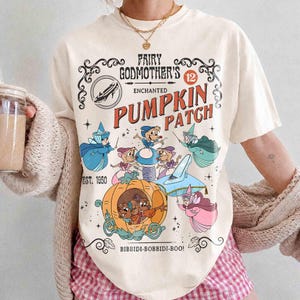 Könnte beinhalten: Ein cremefarbenes T-Shirt mit einem Grafikdruck, der den Text Fairy Godmother's Enchanted Pumpkin Patch, Est. 1950, und Bibbidi-Bobbidi-Boo enthält. Das Design zeigt Illustrationen von Mäusen, einer Kürbiskutsche und einem gläsernen Schuh.