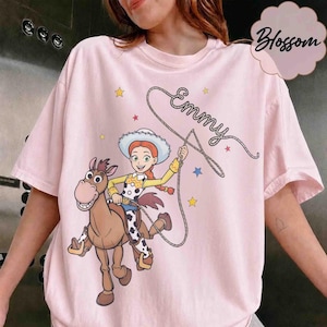 May include: A light pink t-shirt featuring a graphic of a cartoon cowgirl riding a brown horse. The cowgirl holds a lasso that forms the cursive text "Emmy" above her. The design includes small yellow, blue, and red stars scattered around.