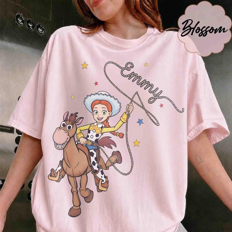May include: A light pink t-shirt featuring a graphic of a cartoon cowgirl riding a brown horse. The cowgirl holds a lasso that forms the cursive text "Emmy" above her. The design includes small yellow, blue, and red stars scattered around.