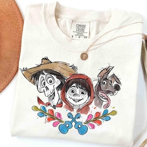 May include: Cream-colored t-shirt featuring a colorful illustration of characters from the animated movie Coco. The design includes a skeleton, a boy in a red hoodie, a dog, and two guitars. A gold necklace is draped across the shirt.