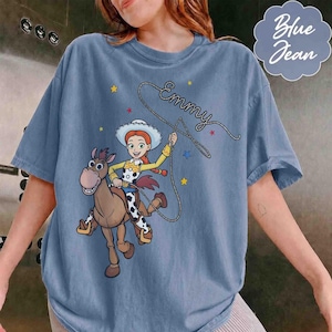 May include: A blue t-shirt featuring a graphic of a cowgirl riding a brown horse. The cowgirl is holding a lasso that forms the cursive text "Emmy" above her head. The design includes small yellow and red stars scattered around the characters.