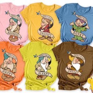 May include: A collection of six colorful t-shirts featuring the seven dwarfs from Snow White. Each shirt displays a different dwarf with a banner that reads "Certified" and their name: Bashful, Grumpy, Dopey, Happy, Sleepy, and Sneezy.
