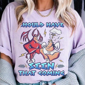 Puede incluir: Una camiseta lila con una ilustración de dibujos animados de un diablo rojo y un ángel, acompañada del texto "SHOULD HAVE SEEN THAT COMING" en azul y blanco. El diseño incorpora rayos y una nube.