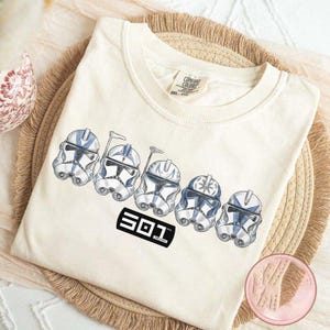 May include: Cream-colored t-shirt featuring a graphic of five blue and white clone trooper helmets. The text "501" is printed in a black rectangle below the helmets. The shirt is folded on a woven mat.