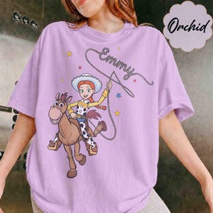 May include: A light purple t-shirt featuring a graphic of a cowgirl riding a brown horse. The cowgirl holds a lasso that forms the cursive text "Emmy" above her head. The design includes small yellow and blue stars scattered around the characters.